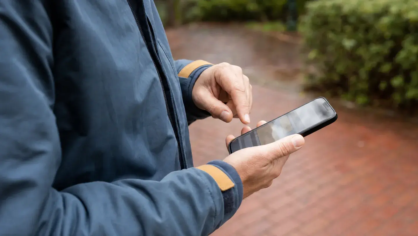 Close-up van handen die buiten een smartphone bedienen, met een blauwgrijze jas en onscherpe bestrating