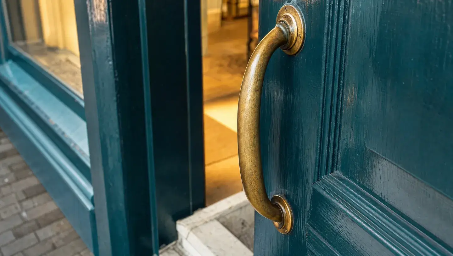 Detail van een blauwgroene houten deur met een messing handgreep en warm verlichte binnenruimte