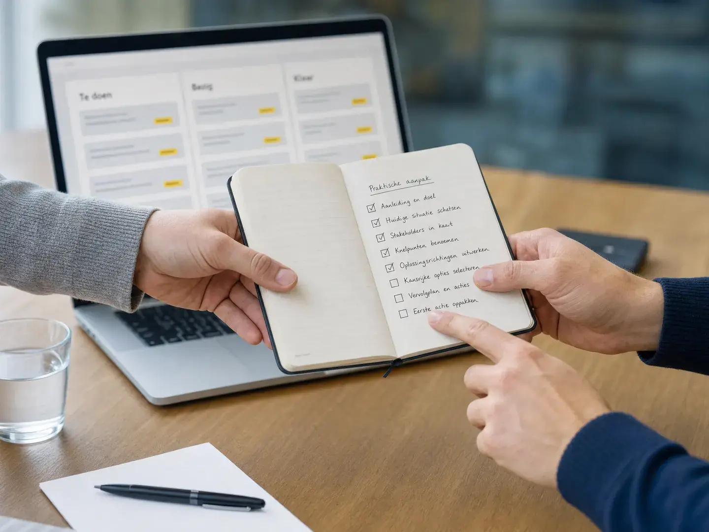 Twee paar handen dragen een notitieboek met werklijst over naast een laptop